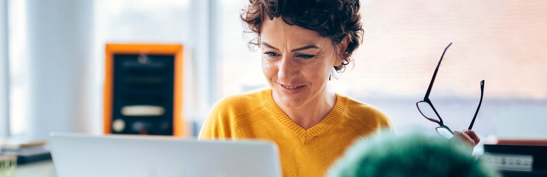 A woman sits and works at her laptop, glasses in hand
